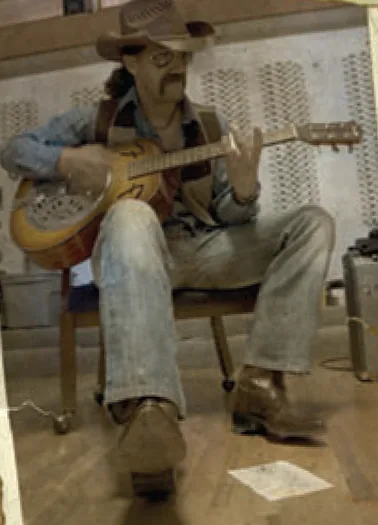 Charlie Bunkum in hat, sunglasses and mustache playing a resonator guitar on a stool at Studio C in Stockton, California