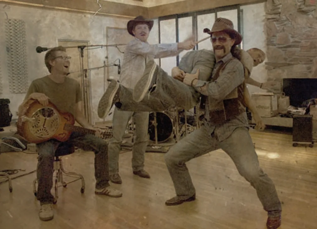 Phil and Charlie Bunkum horsing around mid-kick in the studio with two bandmates, one holding a resonator guitar